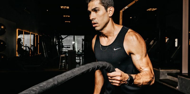 A determined male athlete working out with battle ropes in a dimly lit gym, showcasing strength and fitness.